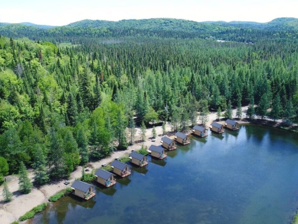 Vue aérienne camping à vendre d'une rangée de cabines flottantes en bois alignées sur la rive d'un lac tranquille, entourées d'une forêt boréale dense au Domaine Lausanne.