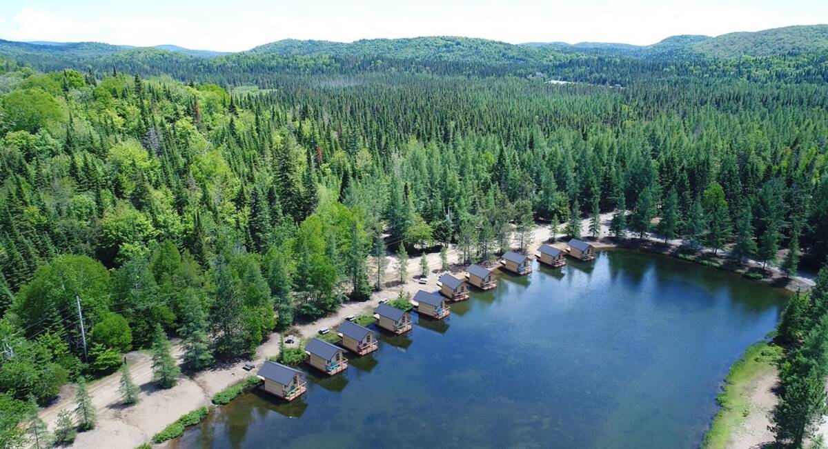 Vue aérienne camping à vendre d'une rangée de cabines flottantes en bois alignées sur la rive d'un lac tranquille, entourées d'une forêt boréale dense au Domaine Lausanne.