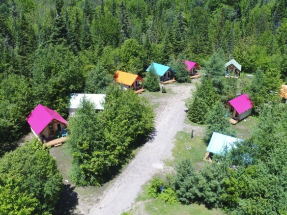 Une vue aérienne montrant un cercle de cabines de prêt-à-camper aux toits colorés (rose, orange, bleu, turquoise) nichées dans une forêt dense au Domaine Lausanne.