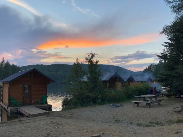 Petits chalets en bois rustiques alignés au bord d'un lac calme au Domaine Lausanne, avec des campeurs profitant d'un repas en plein air sous un ciel de coucher de soleil flamboyant.
