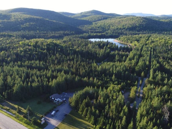 Une vaste vue aérienne du camping à vendre montrant le bâtiment d'accueil et le stationnement bordant une forêt luxuriante qui mène à un lac privé niché dans les montagnes des Laurentides.