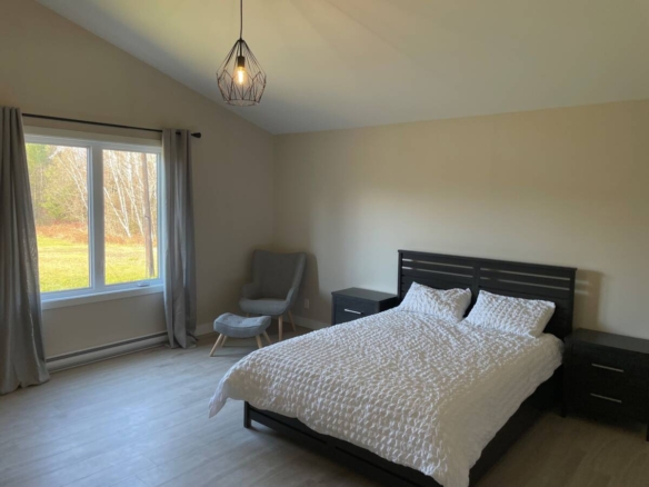 Intérieur d'une chambre minimaliste et lumineuse avec un lit double, un fauteuil gris et une grande fenêtre offrant une vue sur la forêt boréale dans un domaine privé.