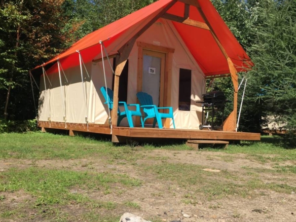 Alt text : Une tente prospecteur de prêt-à-camper avec un toit orange vif sur une plateforme de bois, dotée de deux chaises Adirondack bleues et d'un foyer de pierres au premier plan au Domaine Lausanne.