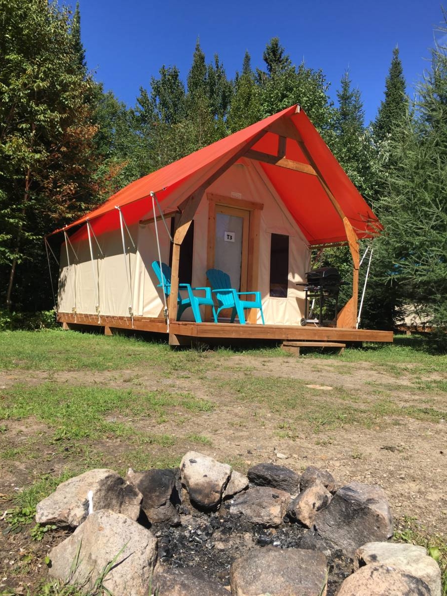 Alt text : Une tente prospecteur de prêt-à-camper avec un toit orange vif sur une plateforme de bois, dotée de deux chaises Adirondack bleues et d'un foyer de pierres au premier plan au Domaine Lausanne.