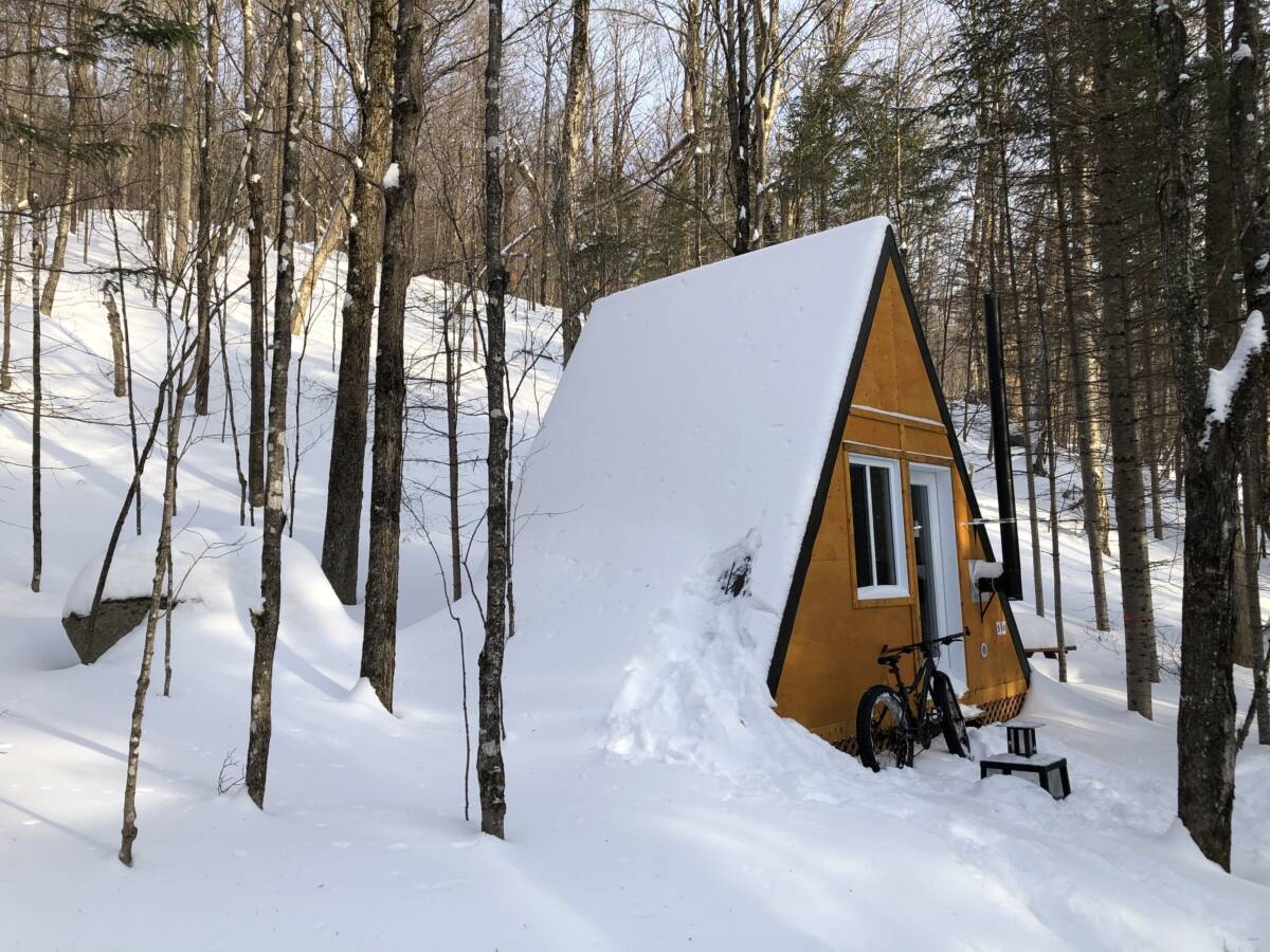 Une cabine en forme de A (A-frame) jaune sous une épaisse couche de neige avec un vélo fatbike stationné devant en forêt au Domaine Lausanne.