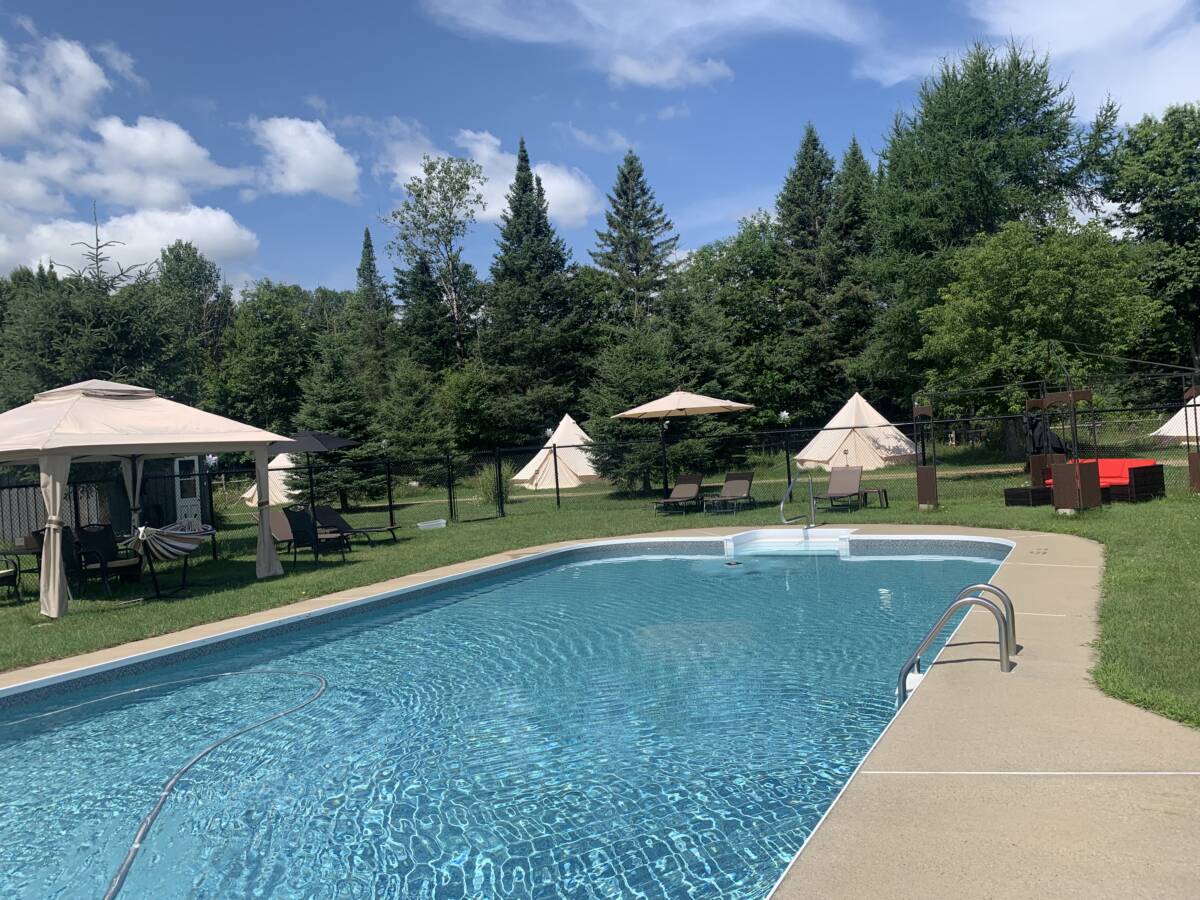 Espace piscine et hébergement glamping – Domaine à vendre Vue de la piscine creusée avec parasols et tentes de glamping sous un ciel bleu au domaine à vendre près de Mont-Tremblant.