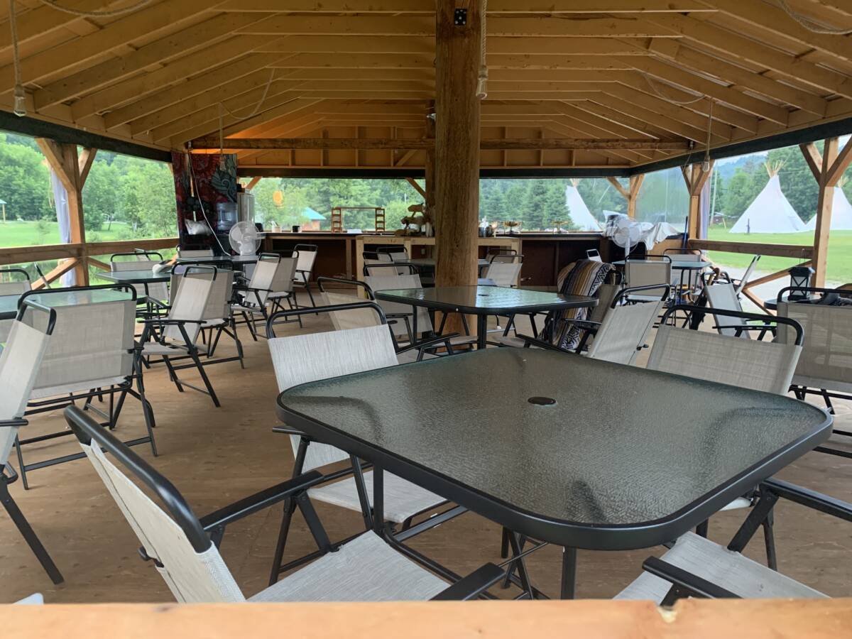 Salle à manger et espace communautaire du centre de ressourcement – Mont-Tremblant Intérieur d'un grand pavillon en bois avec tables en verre et chaises pour groupes sur un domaine de glamping à vendre.