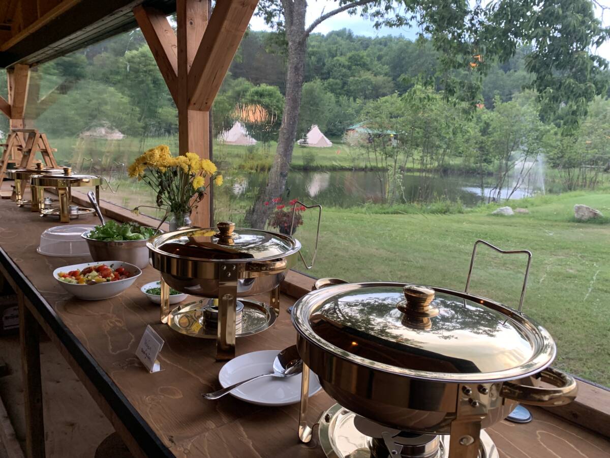 Service de restauration et buffet au centre de ressourcement – Mont-Tremblant Buffet avec plats de service en inox sur une table en bois sous un pavillon ouvert, vue sur l'étang et les tentes de glamping.