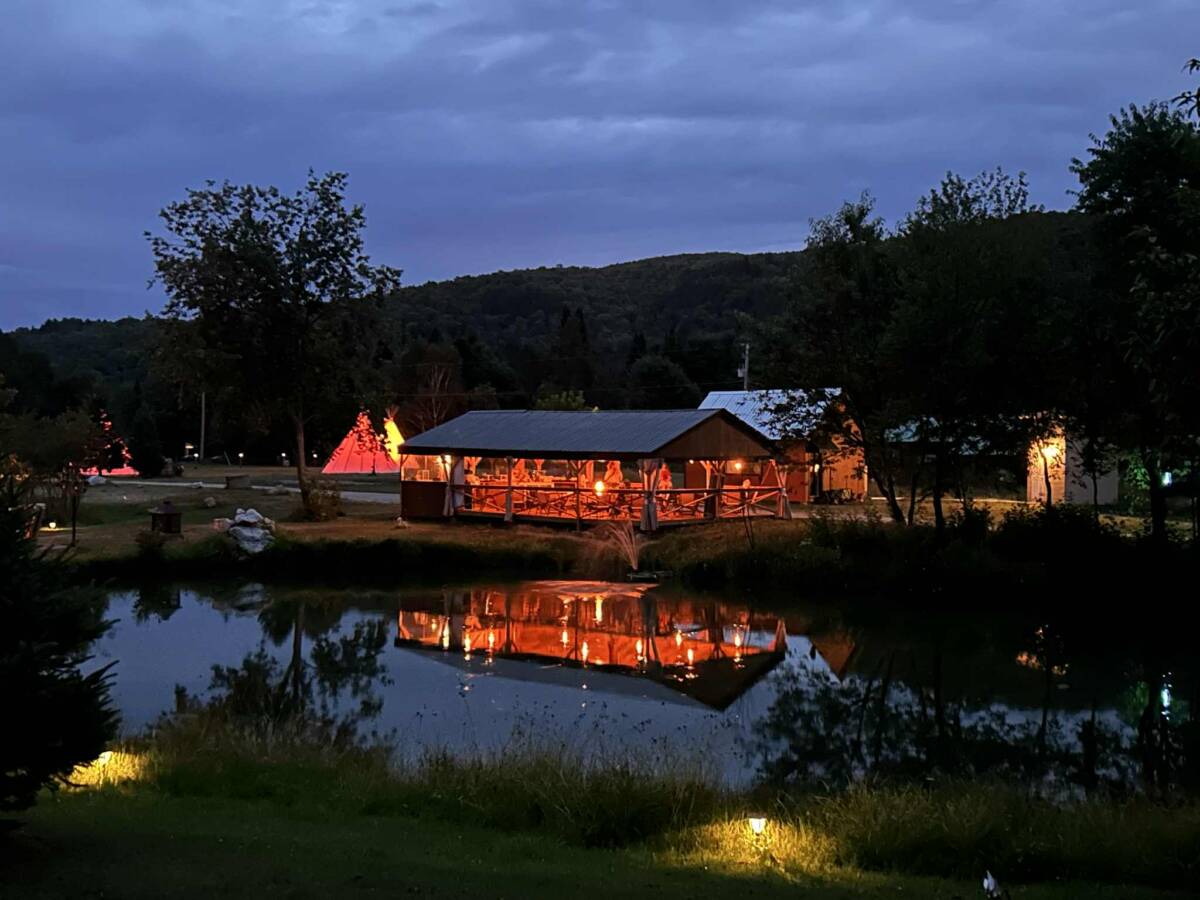 Ambiance nocturne et reflets sur l’étang – Domaine à vendre près de Mont-Tremblant Vue de nuit d'un pavillon en bois illuminé et de tipis se reflétant dans un étang paisible sur un domaine de glamping à vendre.