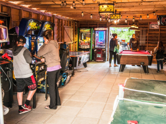 Intérieur de la salle de jeux du Domaine Lausanne montrant des jeunes jouant à des jeux d'arcade, une table de billard et de l'air hockey dans un pavillon rustique en bois.