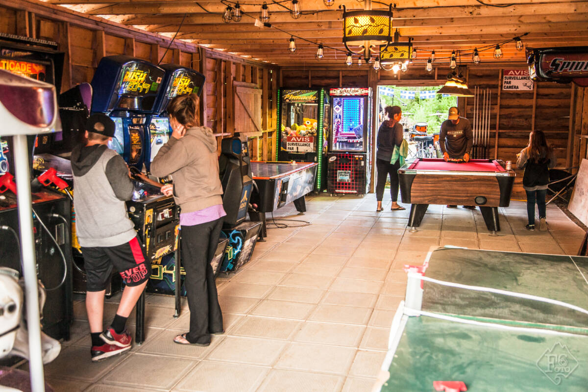 Intérieur de la salle de jeux du Domaine Lausanne montrant des jeunes jouant à des jeux d'arcade, une table de billard et de l'air hockey dans un pavillon rustique en bois.