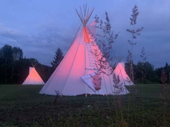 Trois tipis blancs illuminés d'une lueur rose à la tombée de la nuit sur un domaine de glamping à vendre près de Mont-Tremblant.