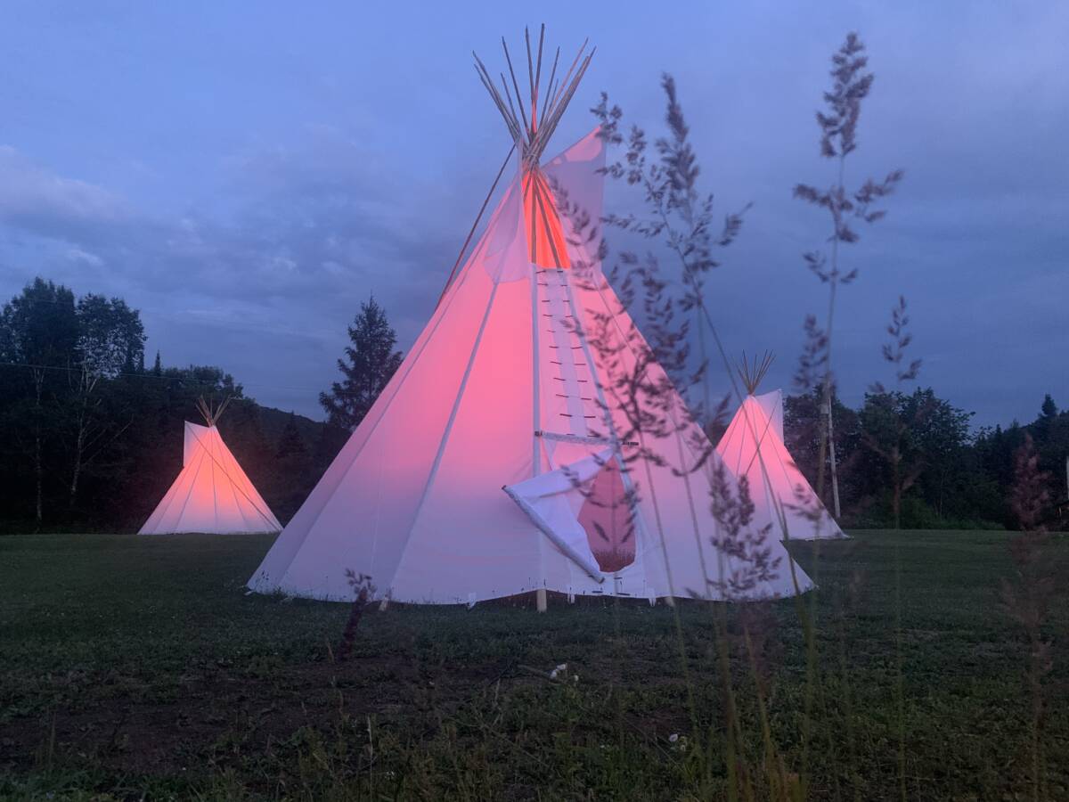 Hébergement insolite en tipi de nuit – Domaine à vendre Trois tipis blancs illuminés d'une lueur rose à la tombée de la nuit sur un domaine de glamping à vendre près de Mont-Tremblant.
