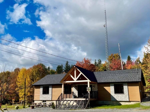 Façade élégante d'un pavillon de réception contemporain en bois et pierre, niché au pied d'une forêt boréale aux couleurs d'automne sous un ciel azur.