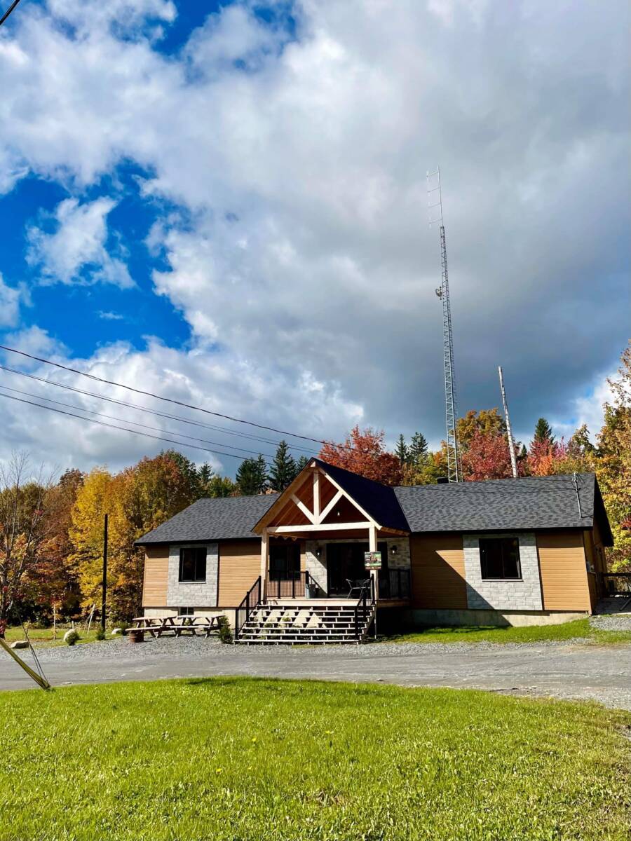 Façade élégante d'un pavillon de réception contemporain en bois et pierre, niché au pied d'une forêt boréale aux couleurs d'automne sous un ciel azur.
