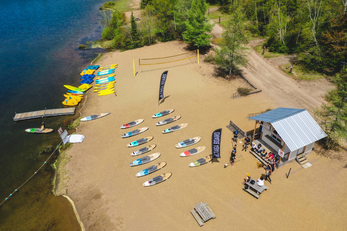 Une vue aérienne plongeante de la plage de sable fin du Domaine Lausanne, montrant des planches à pagaie Taiga Board alignées, un terrain de volleyball et des visiteurs près du centre de location.