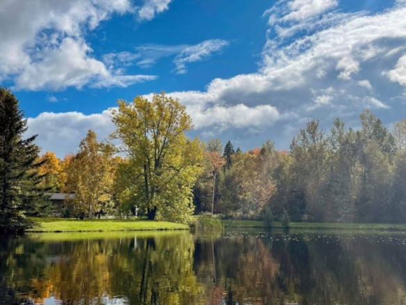 Un lac tranquille au Domaine Confidentiel avec une surface d'eau miroir reflétant des arbres feuillus aux couleurs d'automne jaunes et vertes et un ciel bleu spectaculaire avec des nuages.