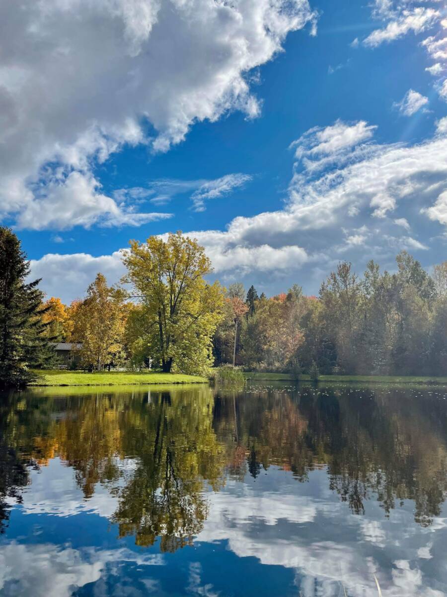 Un lac tranquille au Domaine Confidentiel avec une surface d'eau miroir reflétant des arbres feuillus aux couleurs d'automne jaunes et vertes et un ciel bleu spectaculaire avec des nuages.